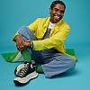 A brown-skinned, smiling Black man with flat twisted hair and wearing a bright yellow and green jacket,  overalls and black Converse All-...