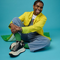 A brown-skinned, smiling Black man with flat twisted hair and wearing a bright yellow and green jacket, overalls and black Converse All-...