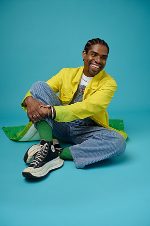 A brown-skinned, smiling Black man with flat twisted hair and wearing a bright yellow and green jacket,  overalls and black Converse All...