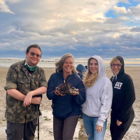 Professor Lisa Jean Moore holds a horseshoe crab with Crispian Thorne '22, Madeline Krol '19, and Anna Hernandez-Krol '15 on Plumb Beach.
