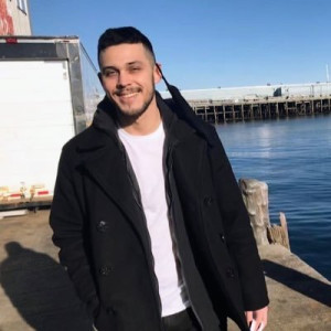 Man in white t-shirt and black jacket smiles with water in background