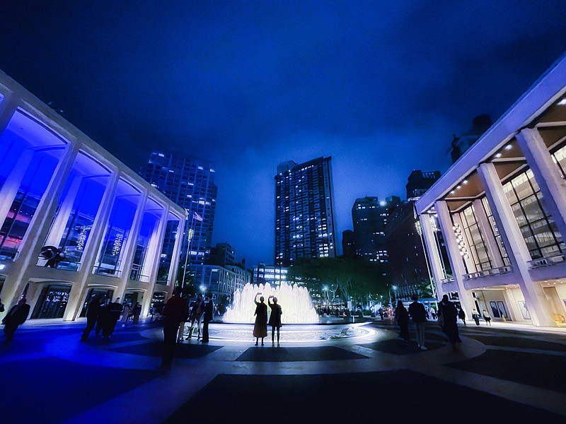 Two people take selfies on a rainy evening at the Lincoln Center fountain.