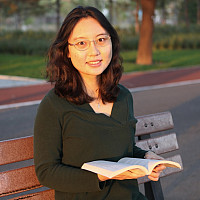 Chuyu Tian wearing dark top and wire rim glasses holding a book open on park bench