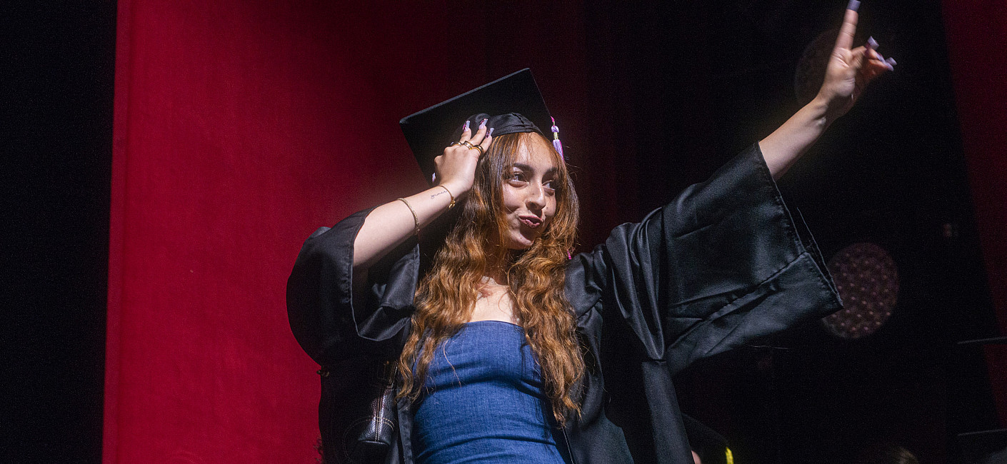 Student in grad cap and gown flashes peace sign to the audience from the stage.