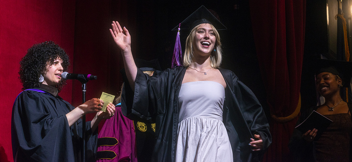 Student in grad cap and gown smiles and waves from the stage.