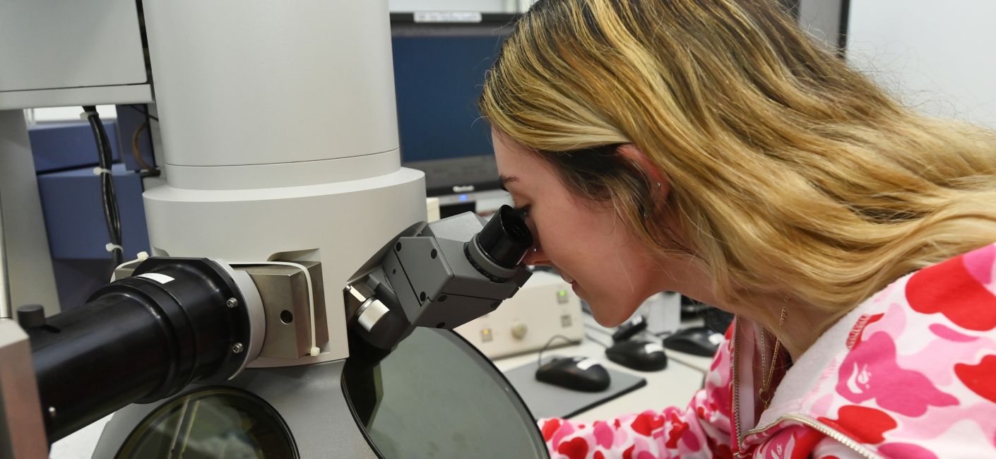 Student with long blond hair peers into a microscope.