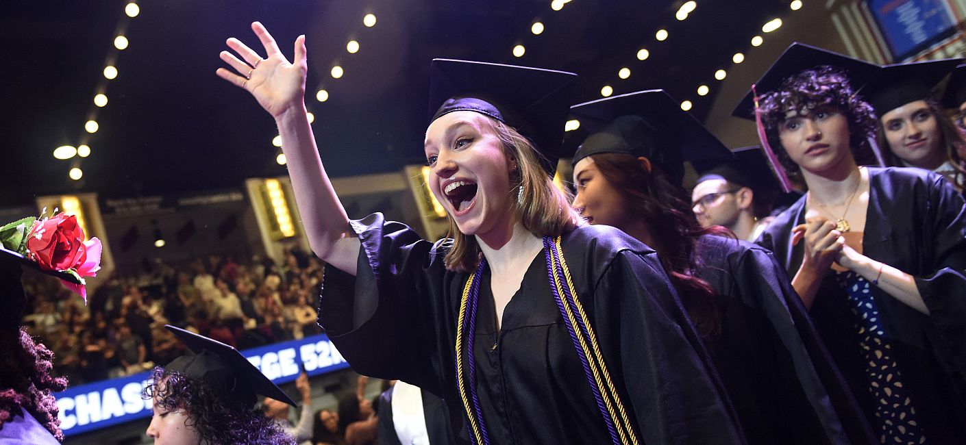 Student in cap and gown in a crowded Westchester County Center smiles and waves.