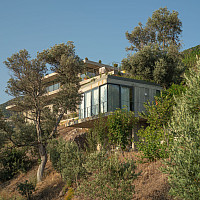 Concrete house on a distant hillside surrounded by trees and greens with a blue sky