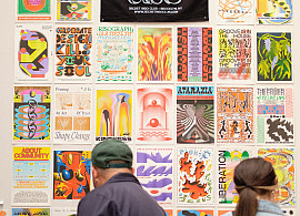 Two people looking at zines on a table, in front of a tableau of risograph-printed posters and a black sign with Secret Riso