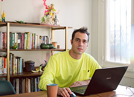 A light complexion person in a yellow shirt with brown hair sits at a computer, holding a mouse. In the background is a book case with bo...
