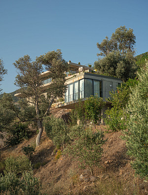 Concrete house on a distant hillside surrounded by trees and greens with a blue sky