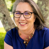 Headshot of Rebecca Albrecht Oling, smiling with greying hair wearing glasses and a chunky necklace