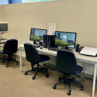 A CCTV, two computer stations, and a braille printer on tables with swivel chairs in the Assistiv...
