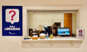 Librarian sitting at Reference Desk window. A poster is on the wall with a big pink question mark and Ask a Librarian written underneath.