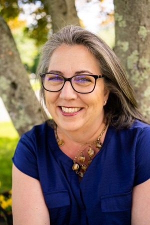 Headshot of Rebecca Albrecht Oling, smiling with greying hair wearing glasses and a chunky necklace